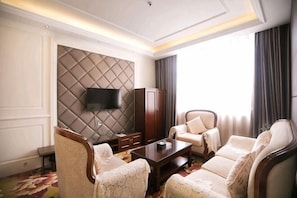 A hotel room with a large flat-screen TV mounted on a padded wall, a sofa set, a wooden coffee table, and a large window with curtains.