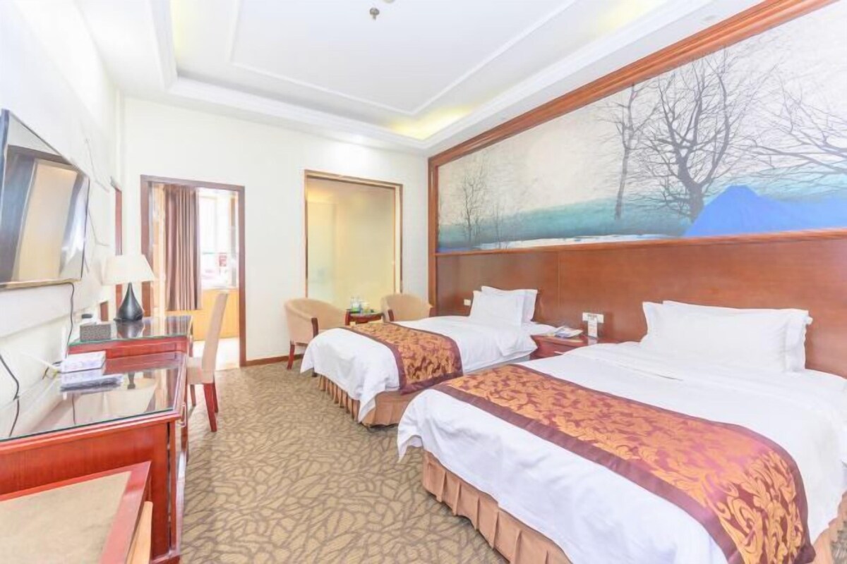 A hotel room with two beds, a desk, a chair, a painting on the wall, and a carpeted floor.