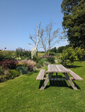 Una mesa de picnic de madera en un jardín con césped, diversas plantas y árboles.