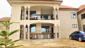 A two-story house with a balcony, columns, and a car parked in front. - Vesgrah Homes (Kumasi)