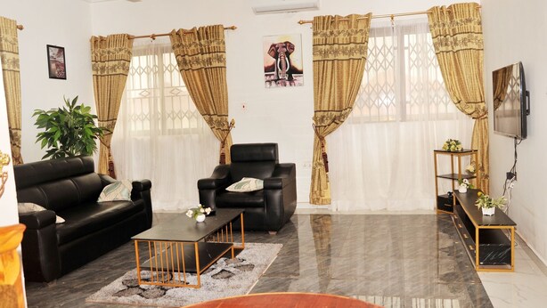 A modern living room with a black leather sofa, a black armchair, a coffee table, and a TV on a stand. There are two large windows with patterned curtains, a potted plant, and a framed picture on the wall. - Vesgrah Homes (Kumasi)