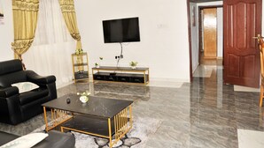 A living room with a black leather sofa, a black coffee table, a flat-screen TV mounted on the wall, and a wooden door leading to another room. - Vesgrah Homes (Kumasi)