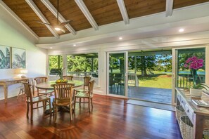 Une pièce spacieuse avec un plafond en bois, de grandes portes-fenêtres coulissantes donnant sur un jardin, une table à manger avec des chaises et un petit bureau avec une lampe.