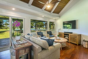 A spacious living room with a large sofa, a round coffee table, and a TV mounted on the wall. - Kealoha - Golf & Lake views, A/C, Pool & Hot Tub (Princeville)