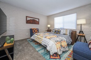 Ein Schlafzimmer mit einem Bett, zwei Nachttischen, einer Lampe, einem Schreibtisch mit einem Kaktus und einem Fenster mit Jalousien.