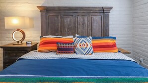 Ein Schlafzimmer mit einem Holz-Kopfteil, einem Bett mit bunter Bettwäsche, einem Nachttisch mit Lampe und einem Spiegel.