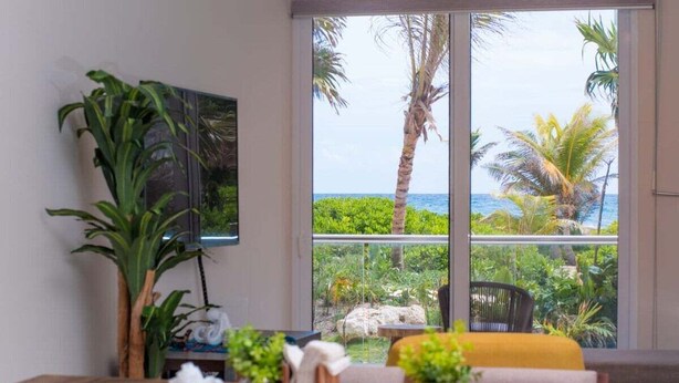 Une pièce dotée d’une grande fenêtre donnant sur un jardin tropical et l’océan. On y trouve une table à manger avec des chaises, ainsi qu’une petite plante.