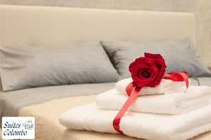 A stack of white towels with a red ribbon and a red rose on a bed with grey pillows. - Suites Colombo (La Spezia)