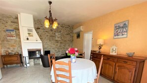 Un comedor con una mesa redonda, sillas de madera, mantel blanco, un jarrón con flores, pared de piedra, chimenea y un aparador de madera.