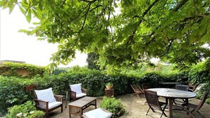 Un patio al aire libre con mobiliario de madera, que incluye sillas y una mesa, rodeado de abundante vegetación y un seto.
