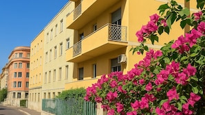 Un edificio amarillo de varios pisos con balcones, cielo azul despejado y flores rosadas al frente.