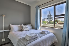 A neatly made bed with white linens and a grey patterned blanket, a bedside lamp, and a window with a view of buildings and trees. - Howard Davis 28B by CTHA (Cape Town)