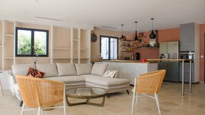 A modern living room with a sectional sofa, two wicker chairs, a glass coffee table, and a kitchen area with a refrigerator, oven, and shelves. - Charming and elegant villa (Tarascon)