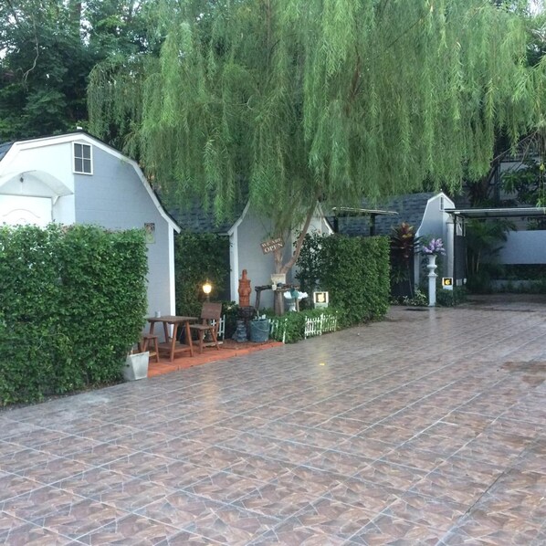 A covered patio area with a tiled floor, surrounded by greenery and a white building.