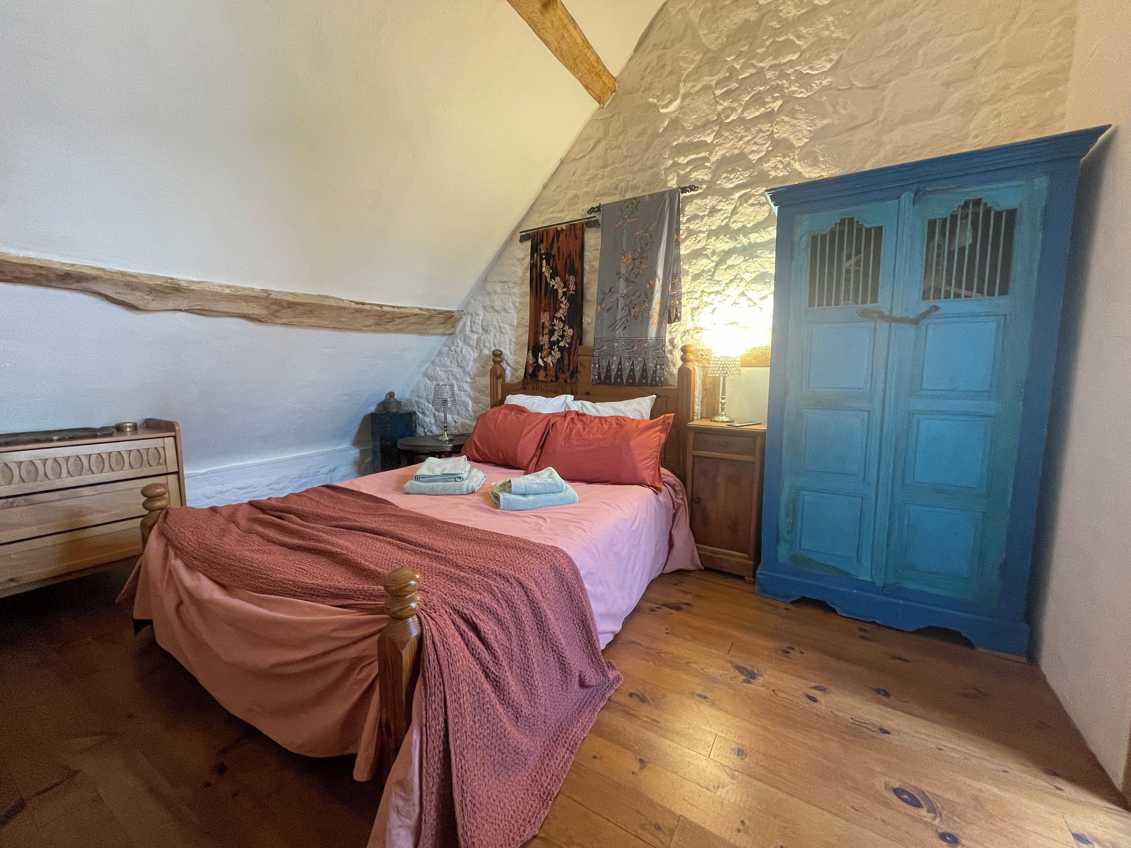 A cozy bedroom with a sloped ceiling, a wooden bed, a nightstand, a blue door, and a small table with a lamp.