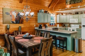 Un intérieur en bois comprenant une table à manger, des chaises, un lustre, un coin cuisine avec des placards et un tableau au mur.
