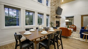 A dining area with a wooden table set for four, surrounded by black chairs, a stone fireplace, and a blue sofa in a modern living room with large windows.