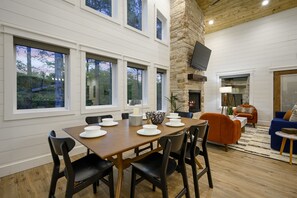 A dining area with a wooden table set for four, surrounded by black chairs, a stone fireplace, and a blue sofa in a modern living room with large windows.