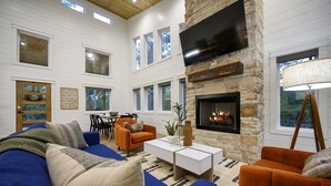 A spacious living room with a stone fireplace, a large flat-screen TV mounted on the wall, and a cozy seating area with a blue sofa and orange armchairs.