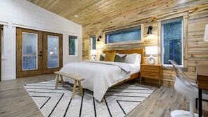 A bedroom with a wooden ceiling, a bed with a white comforter, a bench, a desk with a chair, and a window with a view of trees.