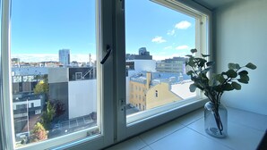 Een kamer met een groot raam dat uitkijkt op een stadsbeeld, een vaas met bloemen op de vensterbank, en een deur met een deurklink.