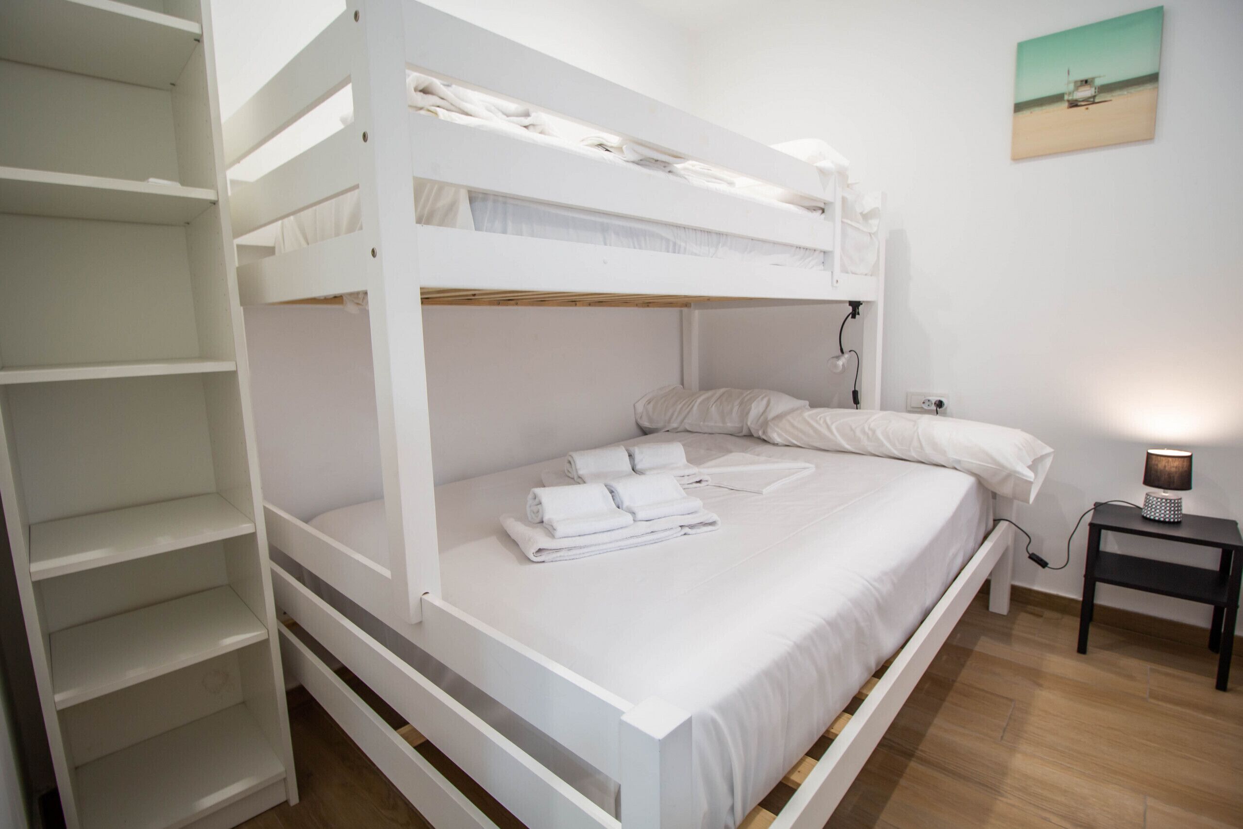 A bunk bed room with a white color scheme, a small bedside table with a lamp, and a painting on the wall.
