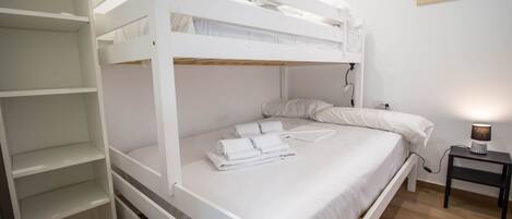 A bunk bed room with a white color scheme, a small bedside table with a lamp, and a painting on the wall.
