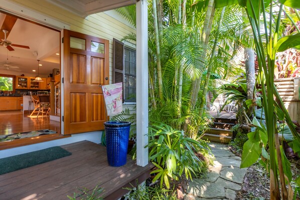 A wooden porch with a door, a blue trash can, and a stone pathway surrounded by tropical plants.