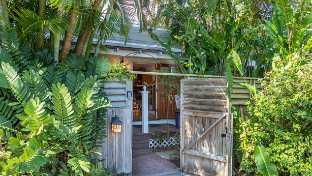 A wooden gate leading to a house with a porch, surrounded by lush greenery.