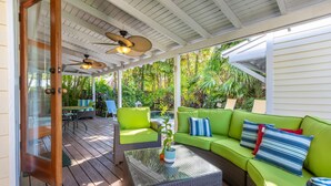 A covered outdoor seating area with wicker furniture, a glass coffee table, and a ceiling fan.