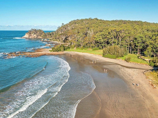 A coastal landscape with a sandy beach, a rocky shoreline, and a forested hill.