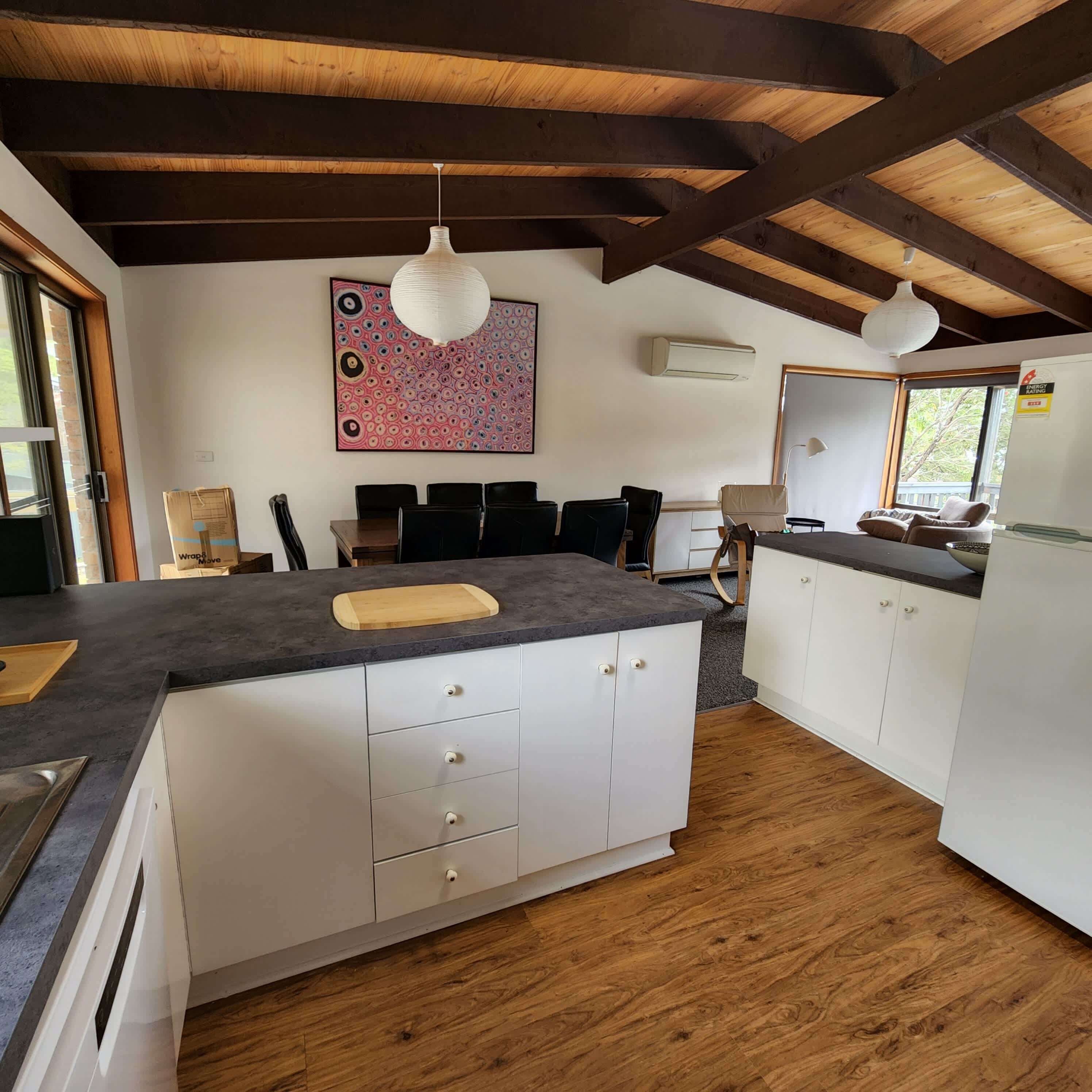A kitchen with a wooden ceiling, white cabinets, a central island with a cutting board, and a colorful abstract painting on the wall.