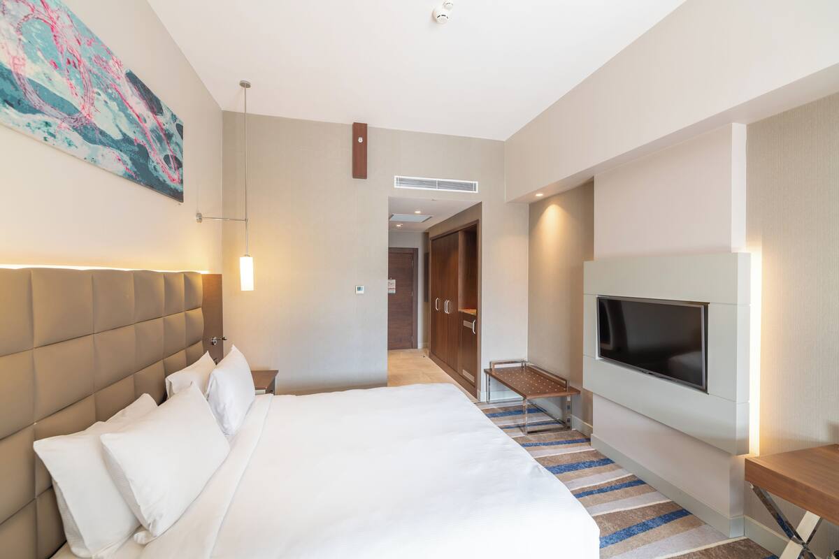 A modern hotel room with a large bed, a flat-screen TV, a painting on the wall, and a hallway leading to a bathroom.