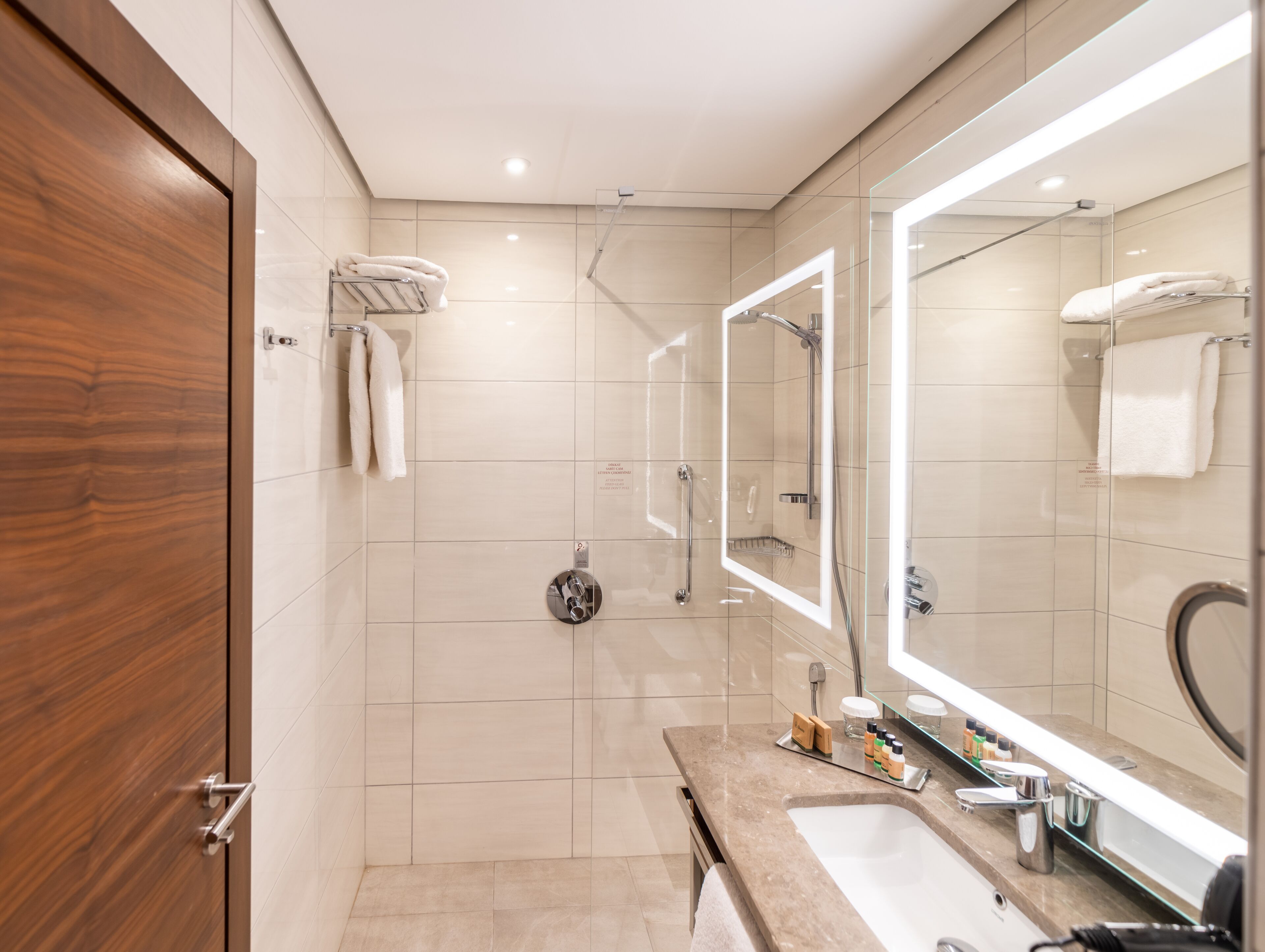 Un baño moderno con puerta de madera, cabina de ducha de vidrio, espejo grande y tocador con artículos de aseo.