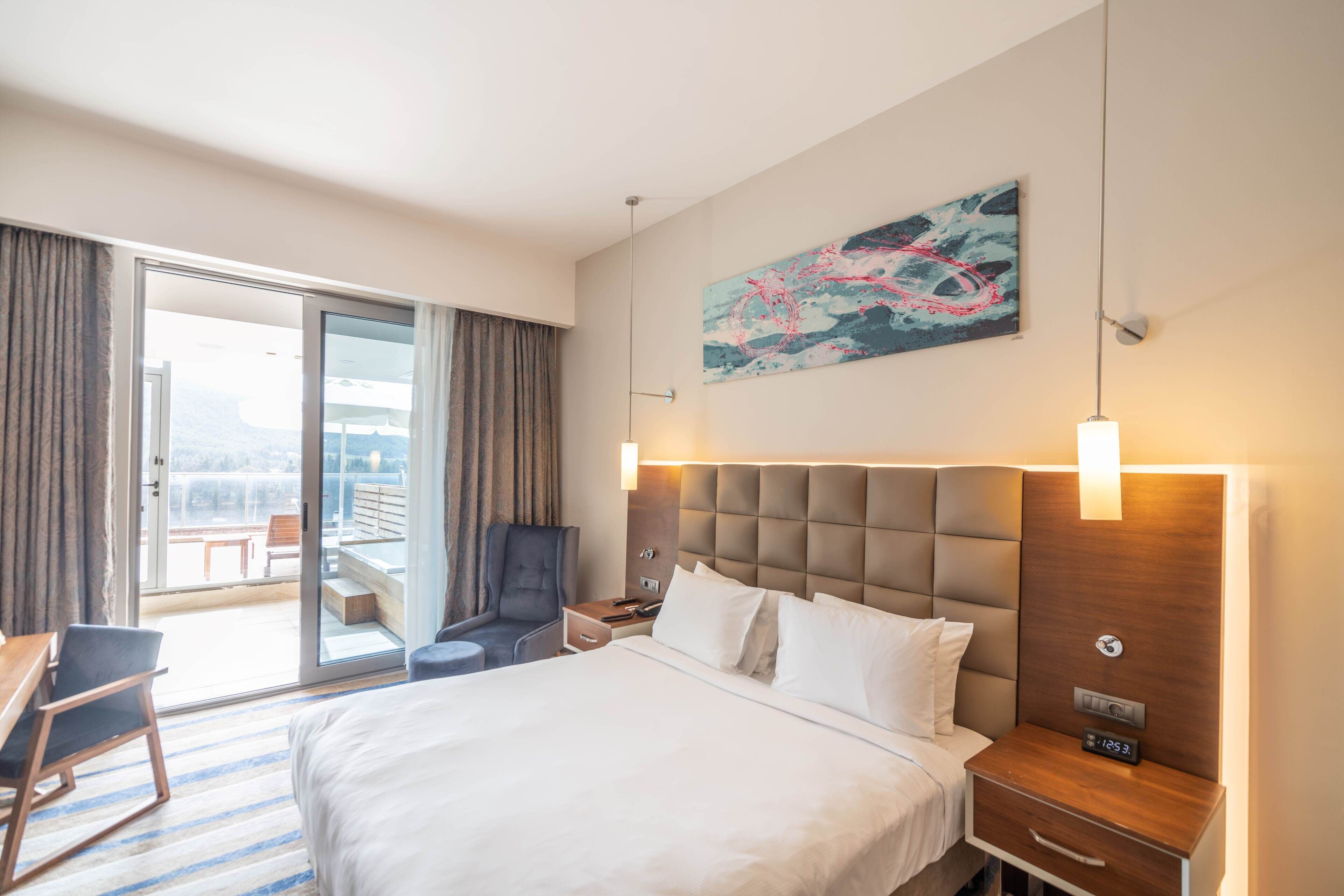 A hotel room with a large bed, a desk, a chair, a nightstand, and a painting on the wall.