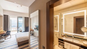 Una habitación de hotel con una cama grande, un escritorio con silla, un baño con espejo y ducha.