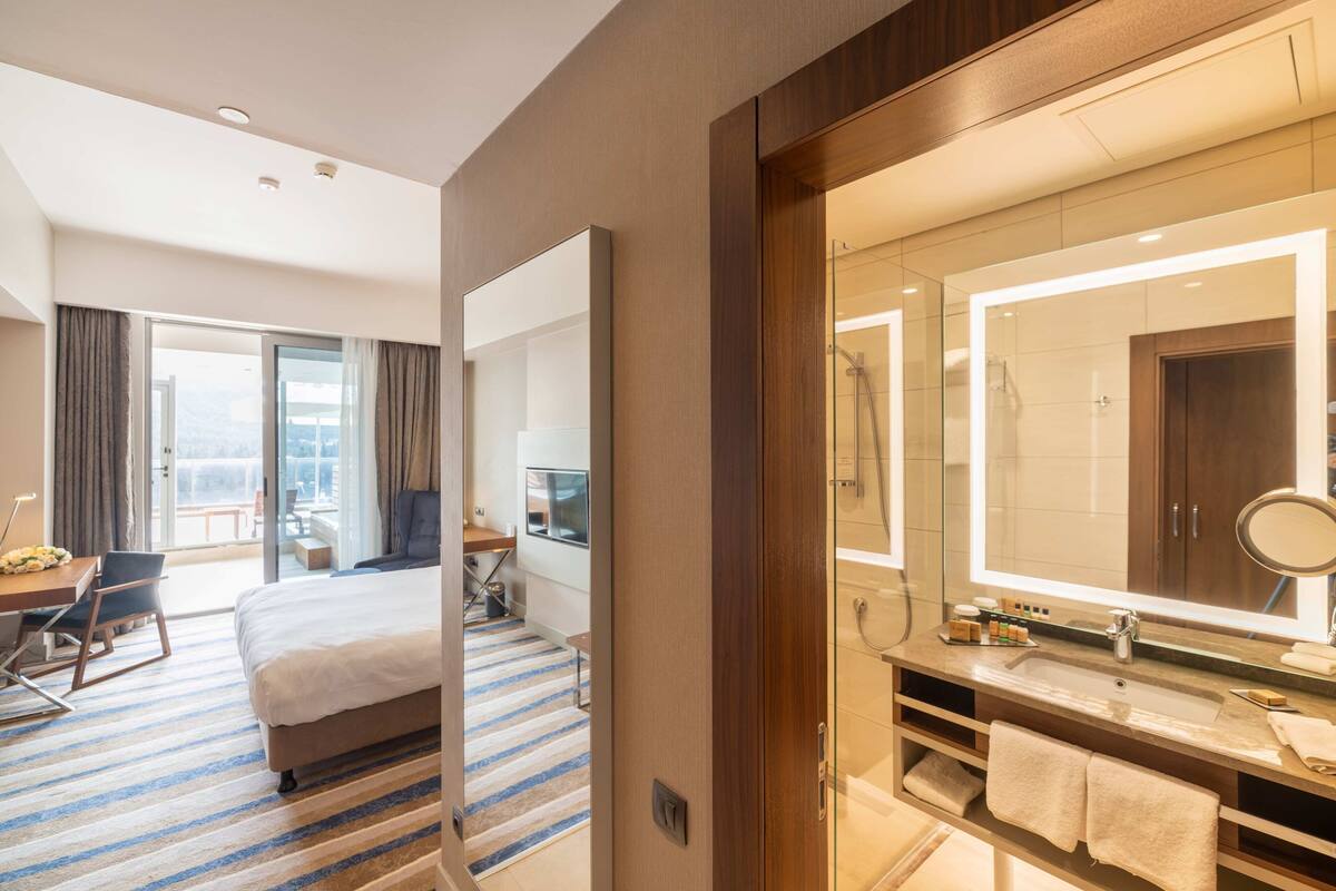 A hotel room with a large bed, a desk with a chair, a bathroom with a mirror, and a shower.