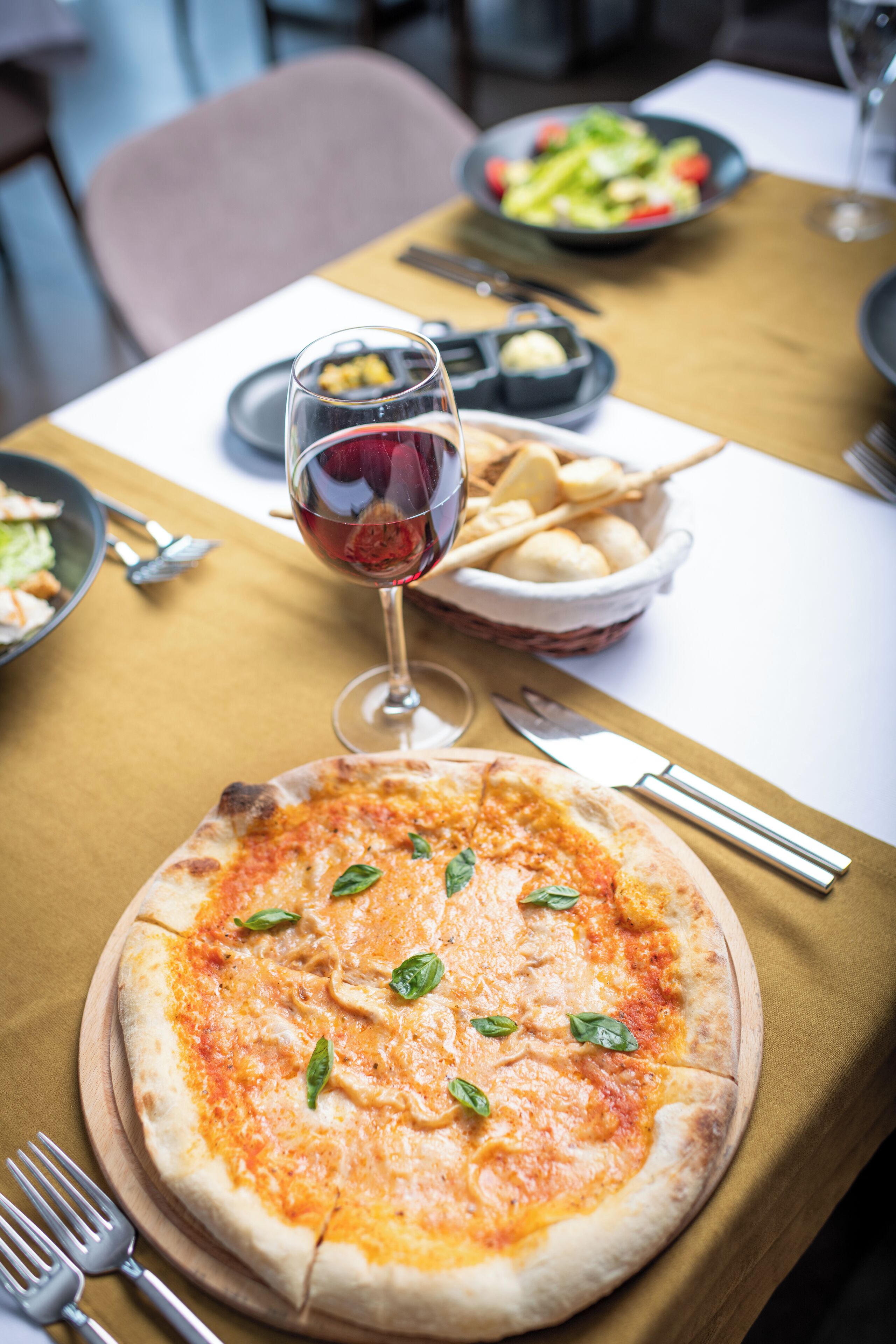 A pizza on a wooden board with a glass of red wine and a basket of breadsticks on a table.
