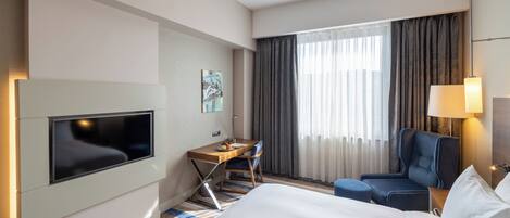 Ein Hotelzimmer mit einem Bett, einem Schreibtisch mit Stuhl, einem Fernseher und einem Fenster mit Vorhängen.