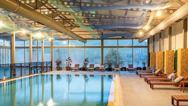 Una piscina interior con paredes de vidrio transparente, sillones de madera para relajarse y vistas de la ciudad a través de amplias ventanas.