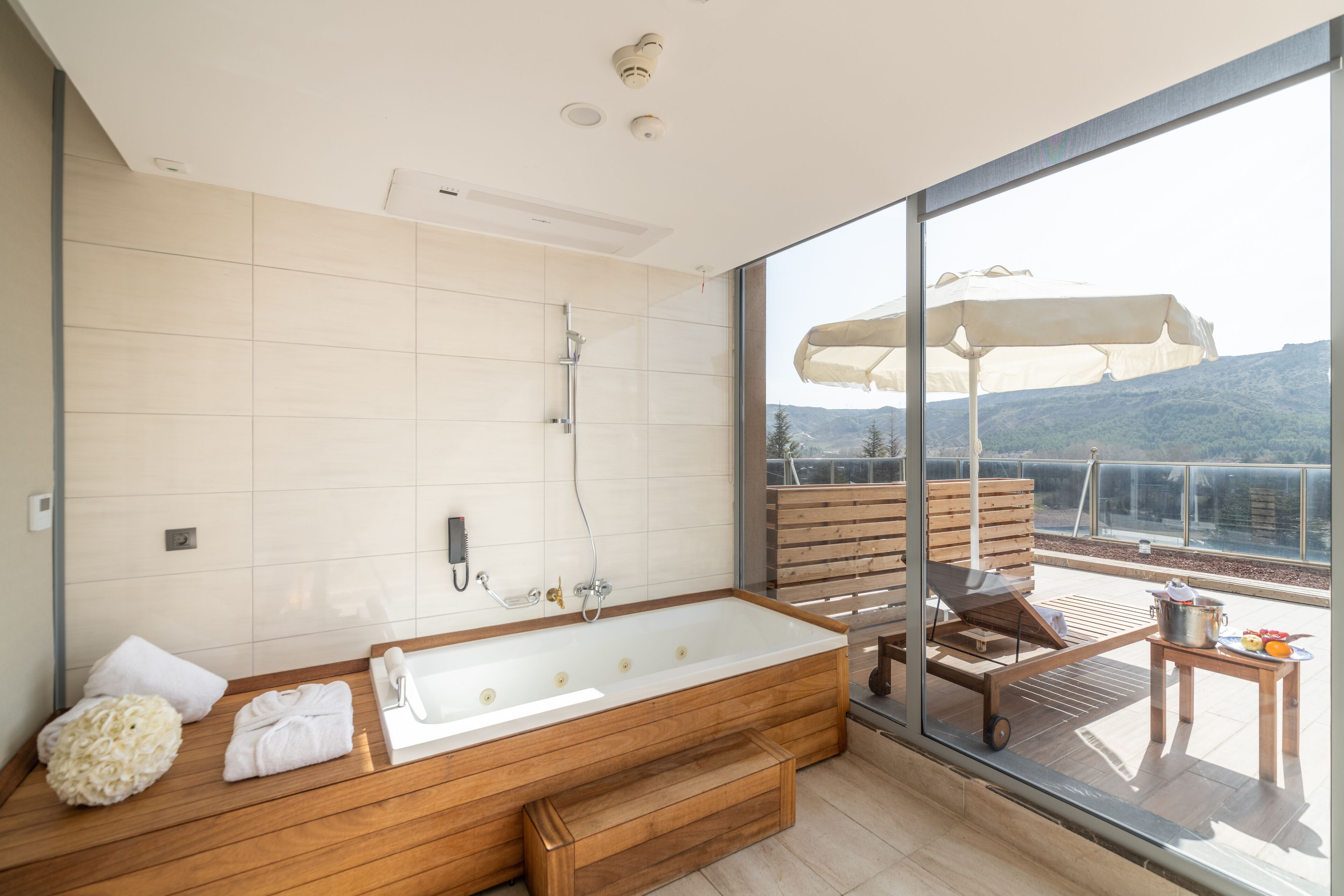 A modern bathroom with a large bathtub, wooden bench, and a view of a balcony with outdoor furniture.