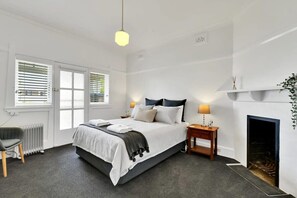 A bedroom with a bed, bedside tables, a radiator, a chair, a fireplace, and a window with blinds.