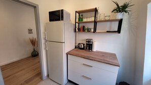 Une cuisine compacte avec un réfrigérateur blanc, un four à micro-ondes, une machine à café et un plan de travail en bois. Des étagères au-dessus du plan de travail servent à ranger des bocaux et une plante en pot.