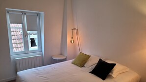 Une chambre avec un grand lit, une petite table ronde, une fenêtre avec des stores et un luminaire fixé au mur.