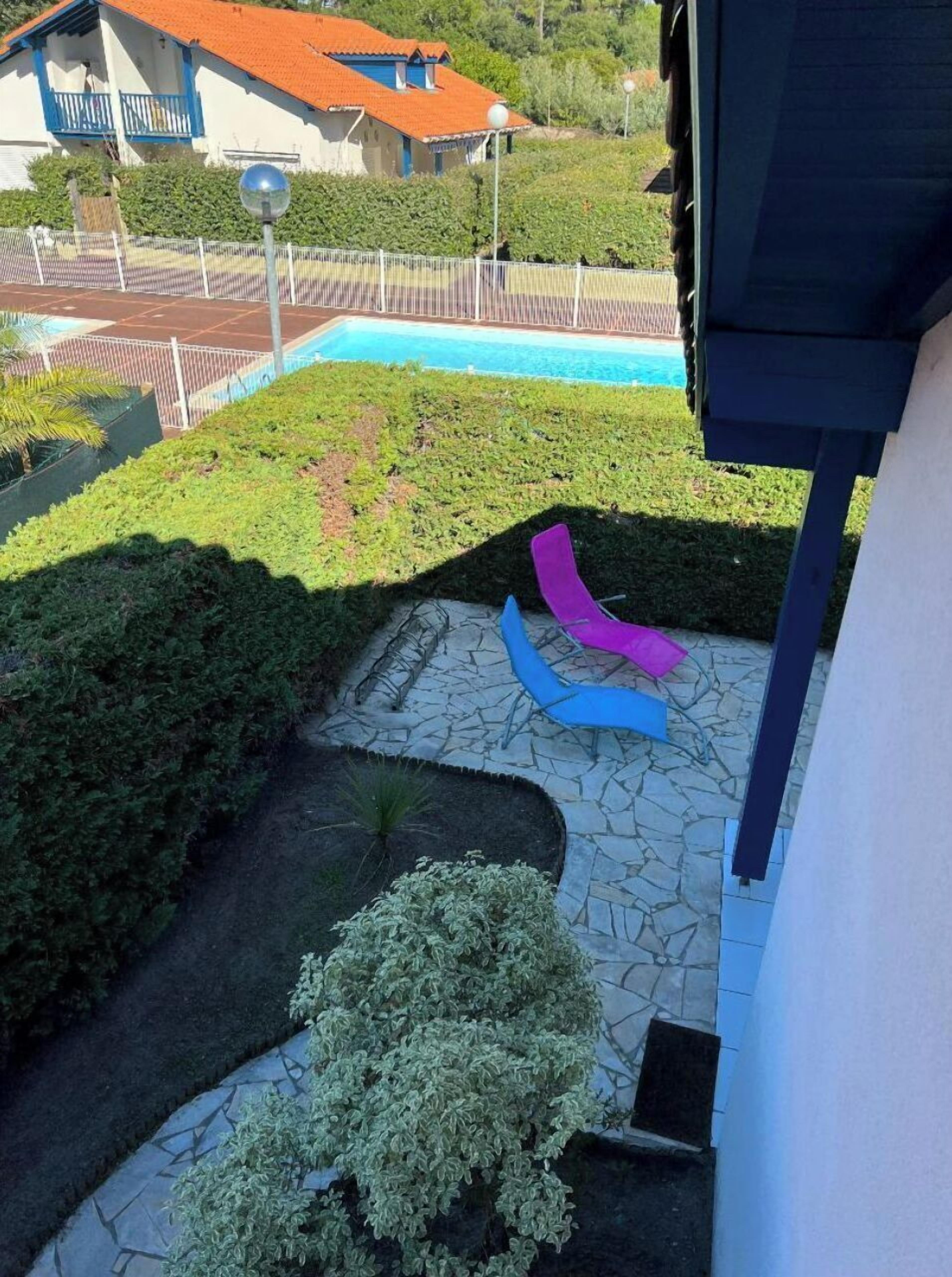 Un espace piscine agrémenté d’un chemin en pierre, d’un petit jardin et, au fond, d’un bâtiment avec un toit rouge.