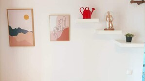 Un mur orné de trois œuvres d’art encadrées, un arrosoir rouge, une petite statue et une plante en pot.
