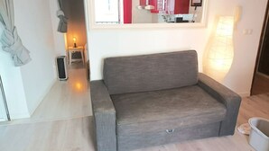 Un salon moderne avec un canapé gris, une petite table avec une lampe et une fenêtre donnant sur un immeuble rouge.