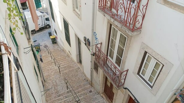 Une ruelle étroite entre deux bâtiments, avec un chemin pavé, un camion blanc garé à gauche et une balustrade de balcon rouge à droite.