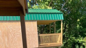 A wooden deck with a green roof, surrounded by dense foliage. - Large Cabin with Stunning View - Fire Pit, Relax & Unwind  (Gallipolis)