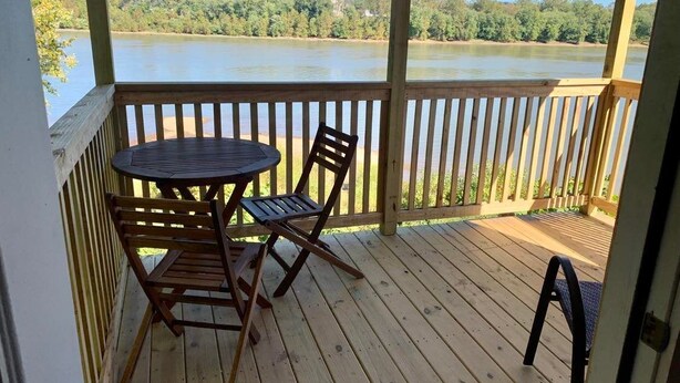 A wooden deck with a table and chairs, overlooking a calm lake with trees and buildings in the background. - Large Cabin with Stunning View - Fire Pit, Relax & Unwind  (Gallipolis)